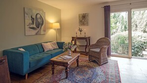 A living room with a blue sofa, a wooden coffee table, a wicker chair, and a large window with a view of trees. - Maison des Cyprès (N°3) (Marcorignan)