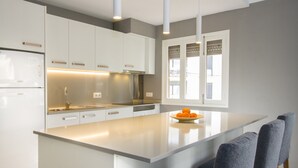 A modern kitchen with white cabinetry, a central island, and a window with blinds. - Apartamentos Ponent MHM (Cala Ratjada)