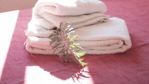 A neatly folded stack of white towels on a bed with a pink bedspread, a decorative plant, and a window with curtains in the background.