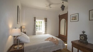 A bedroom with a bed, bedside lamps, a wooden nightstand, a wooden dresser, a ceiling fan, a window with curtains, and a wall clock. - Maison du Néflier (N°2) (Marcorignan)