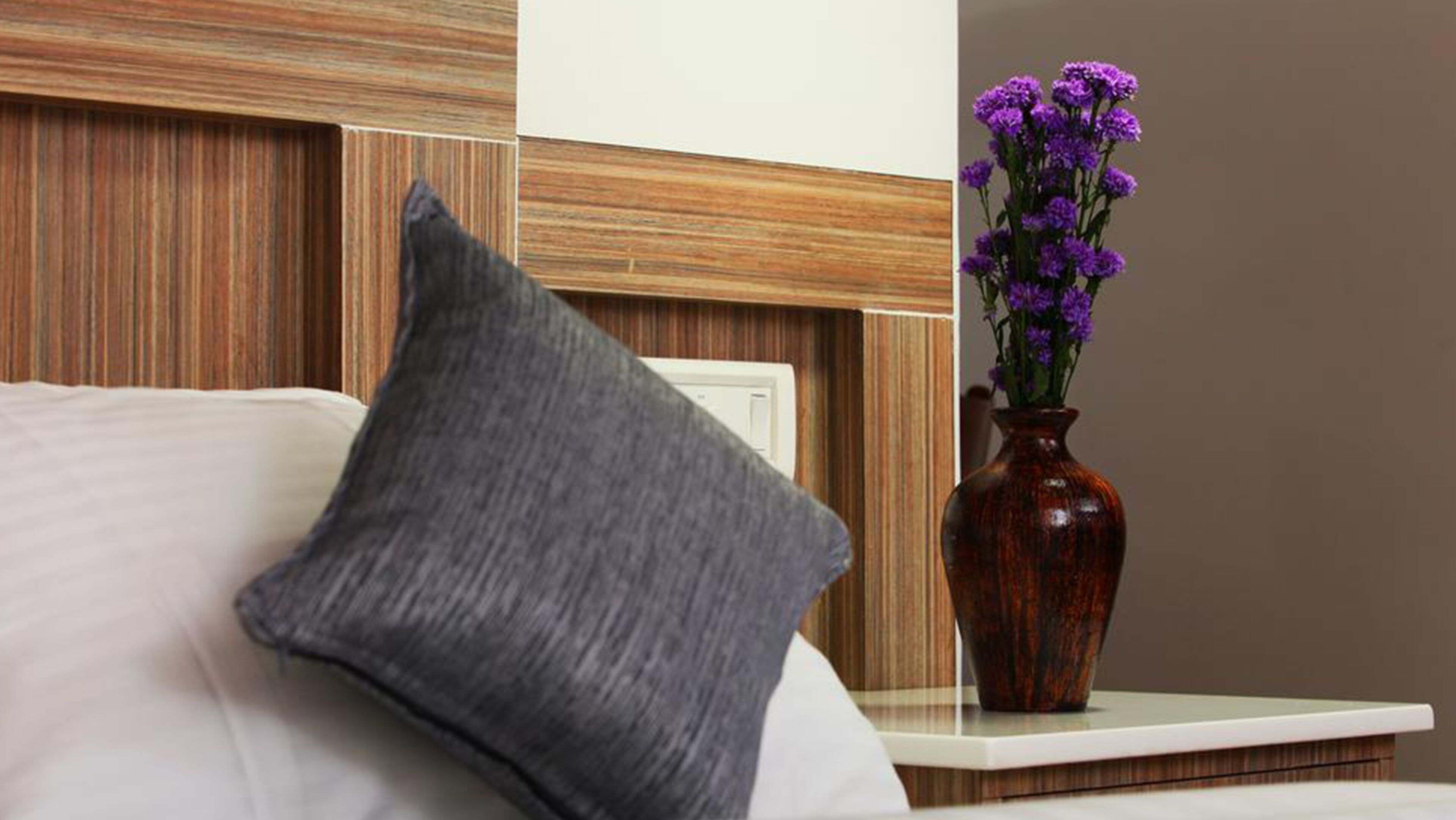 A hotel room with a bed, a grey pillow, and a vase with purple flowers on a nightstand.