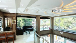 A modern hotel lobby with a high ceiling, a black leather sofa, wooden chairs, a glass coffee table, and large windows offering a view of the city.