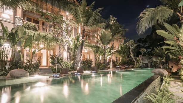 A nighttime view of a pool area with lounge chairs, palm trees, and a building with balconies. - Weda Cita Resort And Spa (Ubud)
