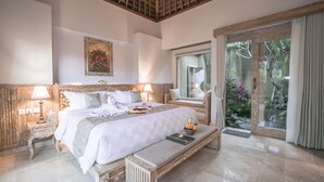 A spacious bedroom with a large bed, wooden headboard, bedside lamps, a bench, and a view of greenery through a glass door. - Weda Cita Resort And Spa (Ubud)