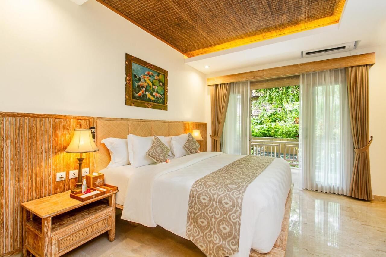 A hotel room with a large bed, a nightstand with a lamp, a painting on the wall, and a view of greenery outside.