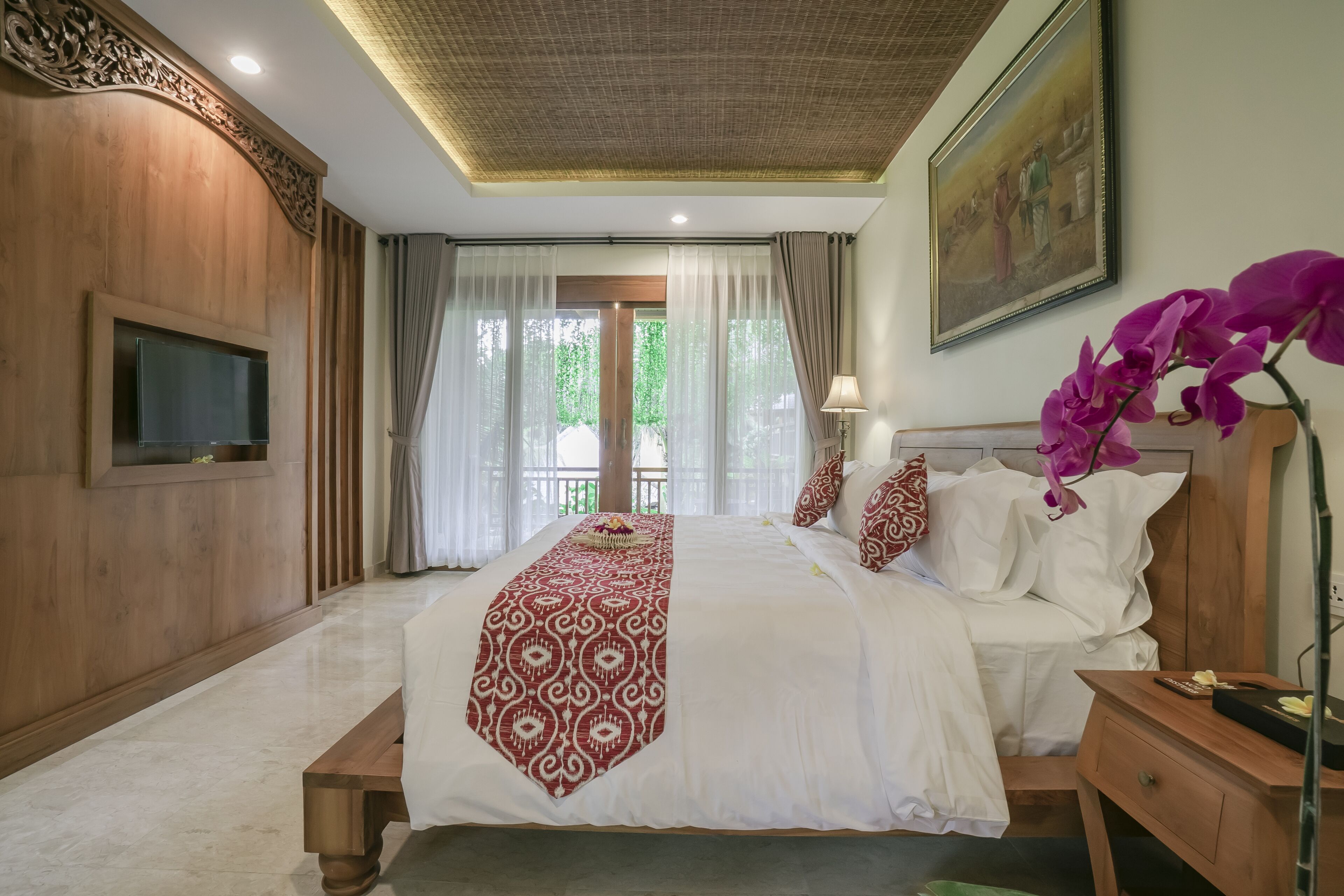 A hotel room with a large bed, wooden paneling, a flat-screen TV, a painting on the wall, and a view of greenery outside.