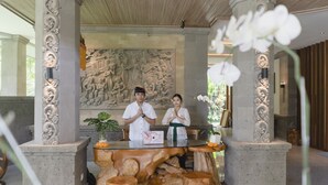A reception area with a central wooden table, two people standing behind it, and a stone wall with carvings in the background. - Weda Cita Resort And Spa (Ubud)