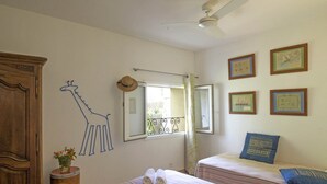A bedroom with two beds, a wooden wardrobe, a ceiling fan, and a window with a view of a balcony. - Maison du Prunier (N°5) (Marcorignan)