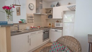 A compact kitchen with white cabinets, a stainless steel sink, a microwave, and a white refrigerator. There is a wicker chair with a floral cushion, a clock on the wall, and a vase with flowers on the counter. - Maison du Prunier (N°5) (Marcorignan)
