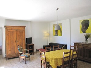 A living room with a wooden cabinet, a television, a sofa, a dining table with chairs, and artwork on the walls.