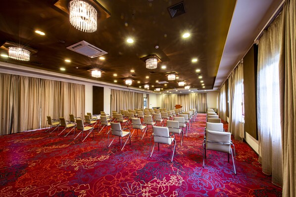 A conference room with rows of chairs, a red carpet, chandeliers, and large windows with curtains.