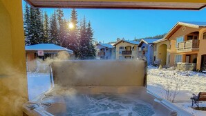 Jacuzzi amb revestiment de fusta, envoltat de terra coberta de neu i edificis al fons.