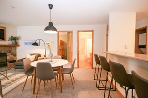 A cozy living room with a fireplace, a round dining table with chairs, a large pendant light, and a kitchen area with bar stools.