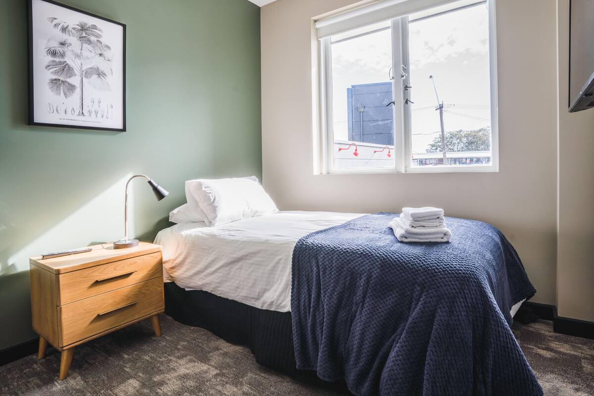 Una camera da letto con un letto, un comodino, una lampada, un'illustrazione botanica incorniciata e una finestra con vista su un edificio.