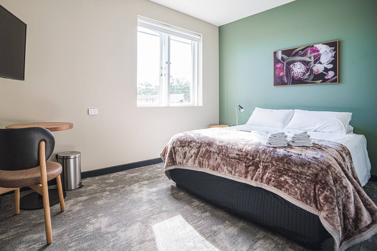 Camera d'albergo con un letto, una sedia, un tavolino, un cestino, una televisione e una finestra.