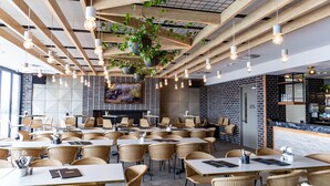 Restaurante moderno com pé-direito alto, vigas aparentes e plantas penduradas. Mesas dispostas em um ambiente amplo, com cadeiras de vime.
