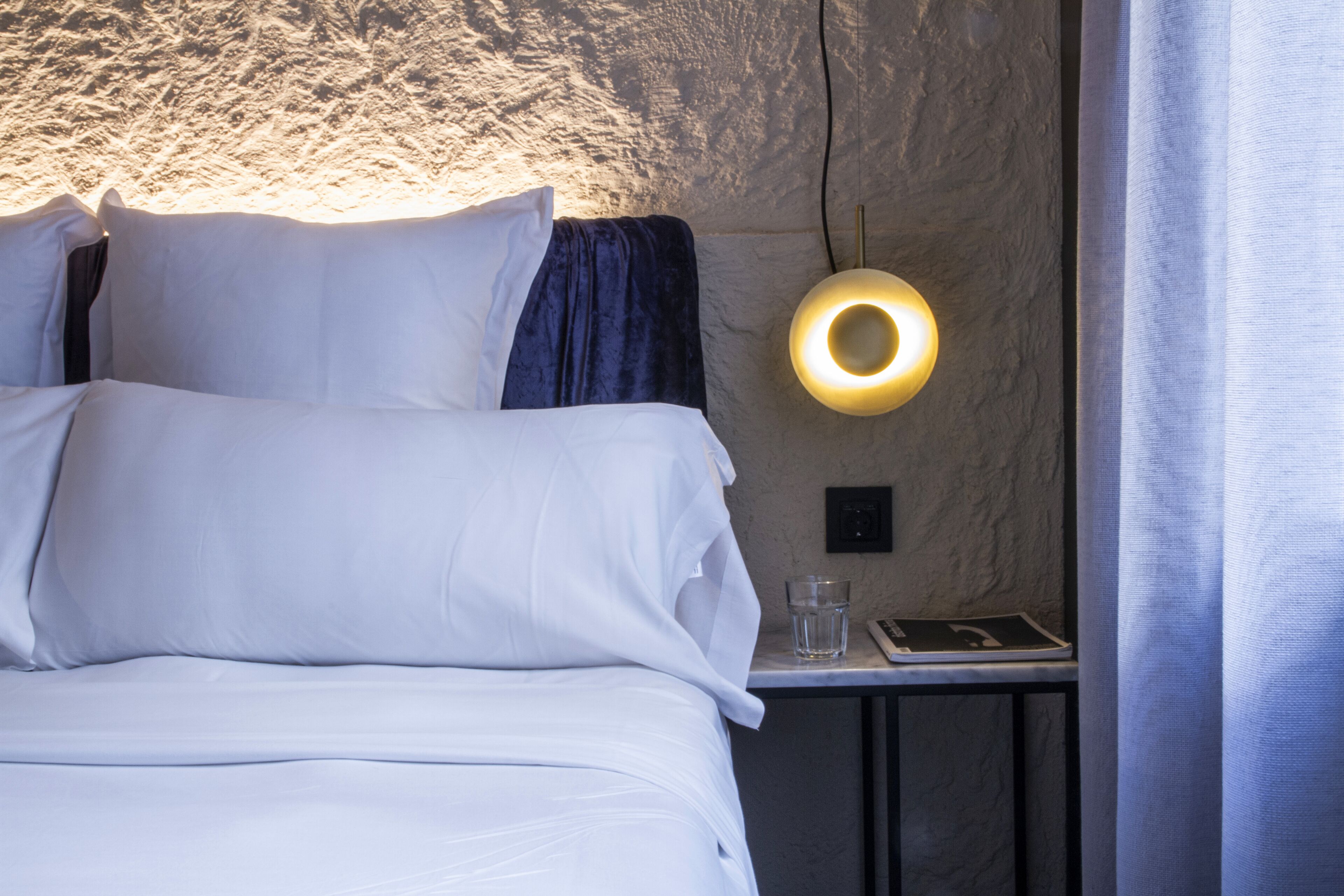 A neatly made bed with white linens, a bedside table with a glass of water and a book, and a wall-mounted light fixture.