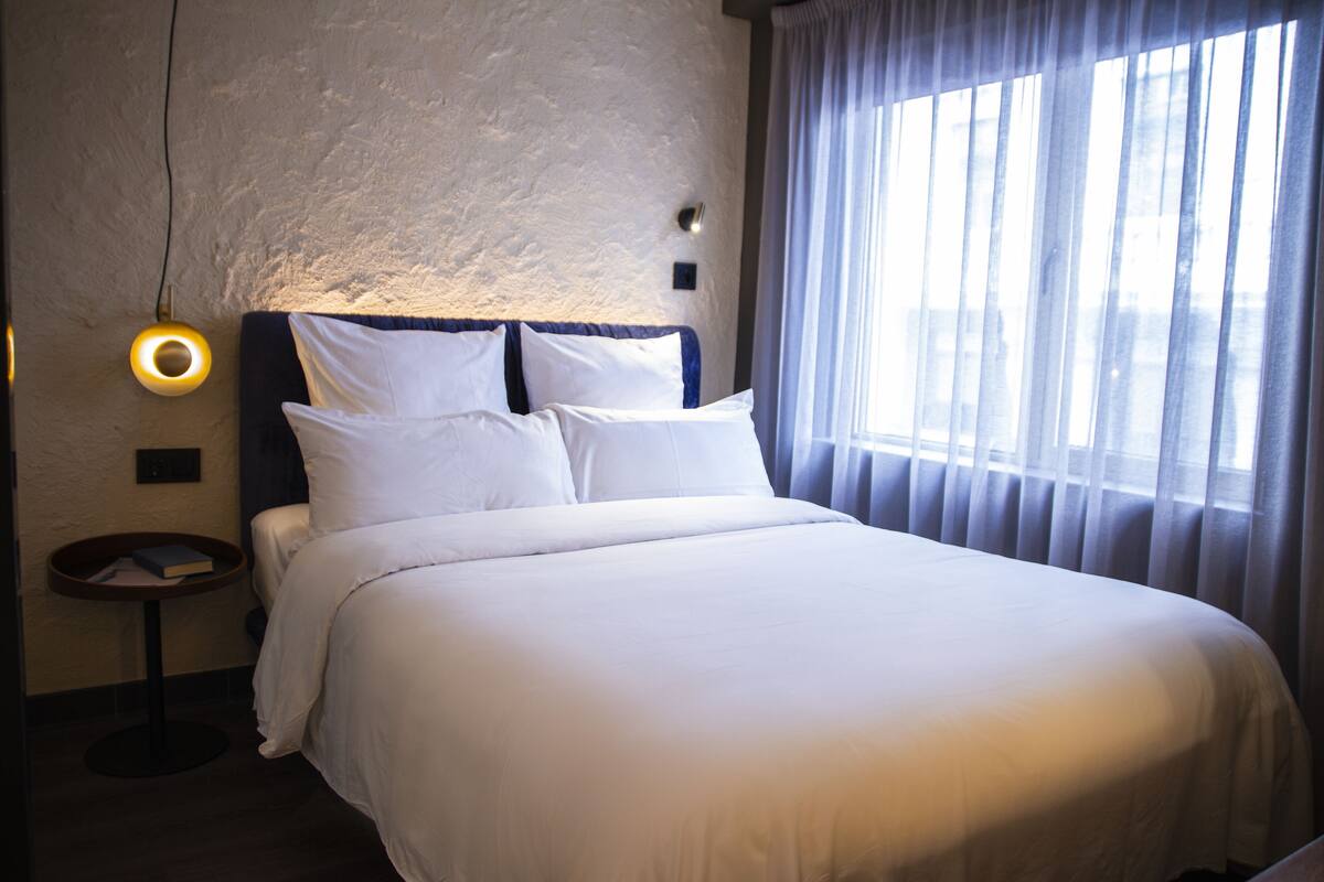 Un lit bien fait, avec du linge de lit blanc, une table de chevet ronde sur laquelle repose un livre, et un luminaire fixé au mur.