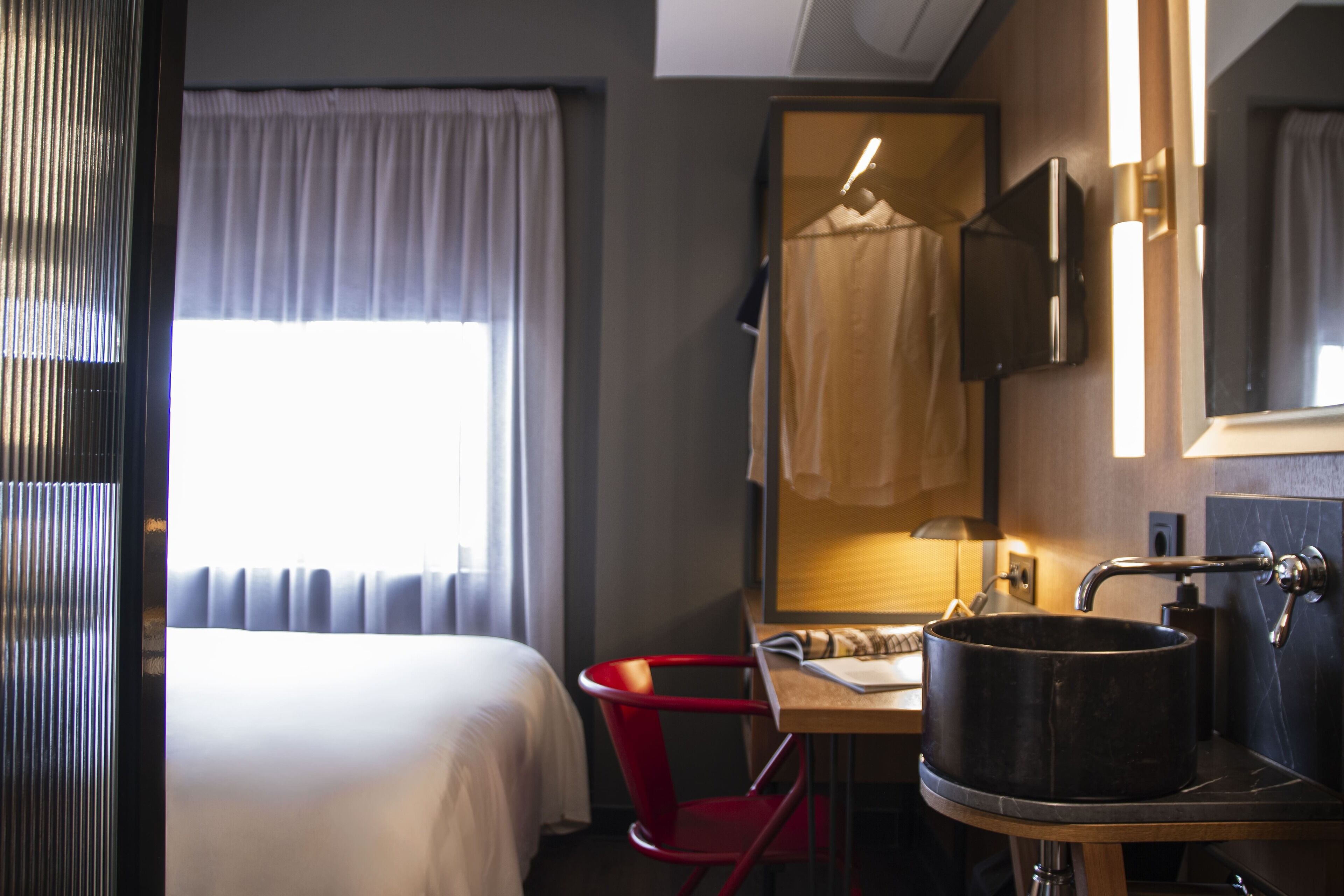 Una habitación de hotel con cama, escritorio con lavamanos, una silla roja y un espejo.