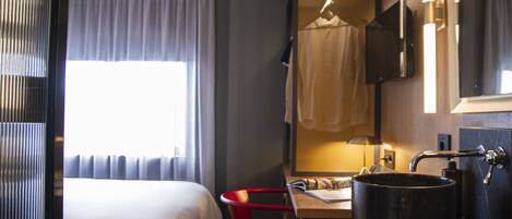 Una habitación de hotel con cama, escritorio con lavamanos, una silla roja y un espejo.