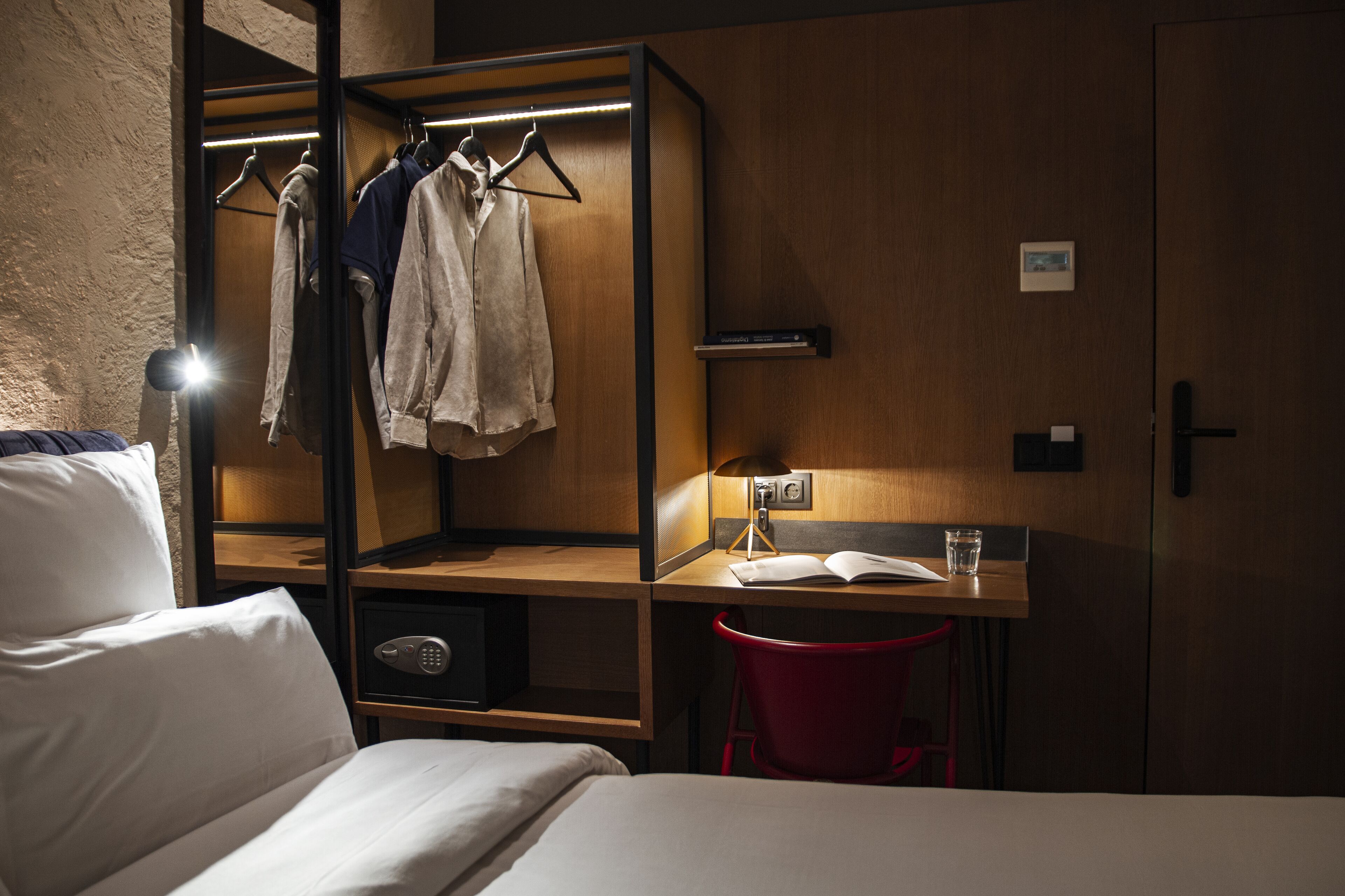 A hotel room with a bed, a desk with a chair, a wardrobe with clothes hanging, and a red trash bin.