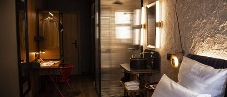 A hotel room with a bed, a desk with a chair, a mirror, and a small table with a kettle.