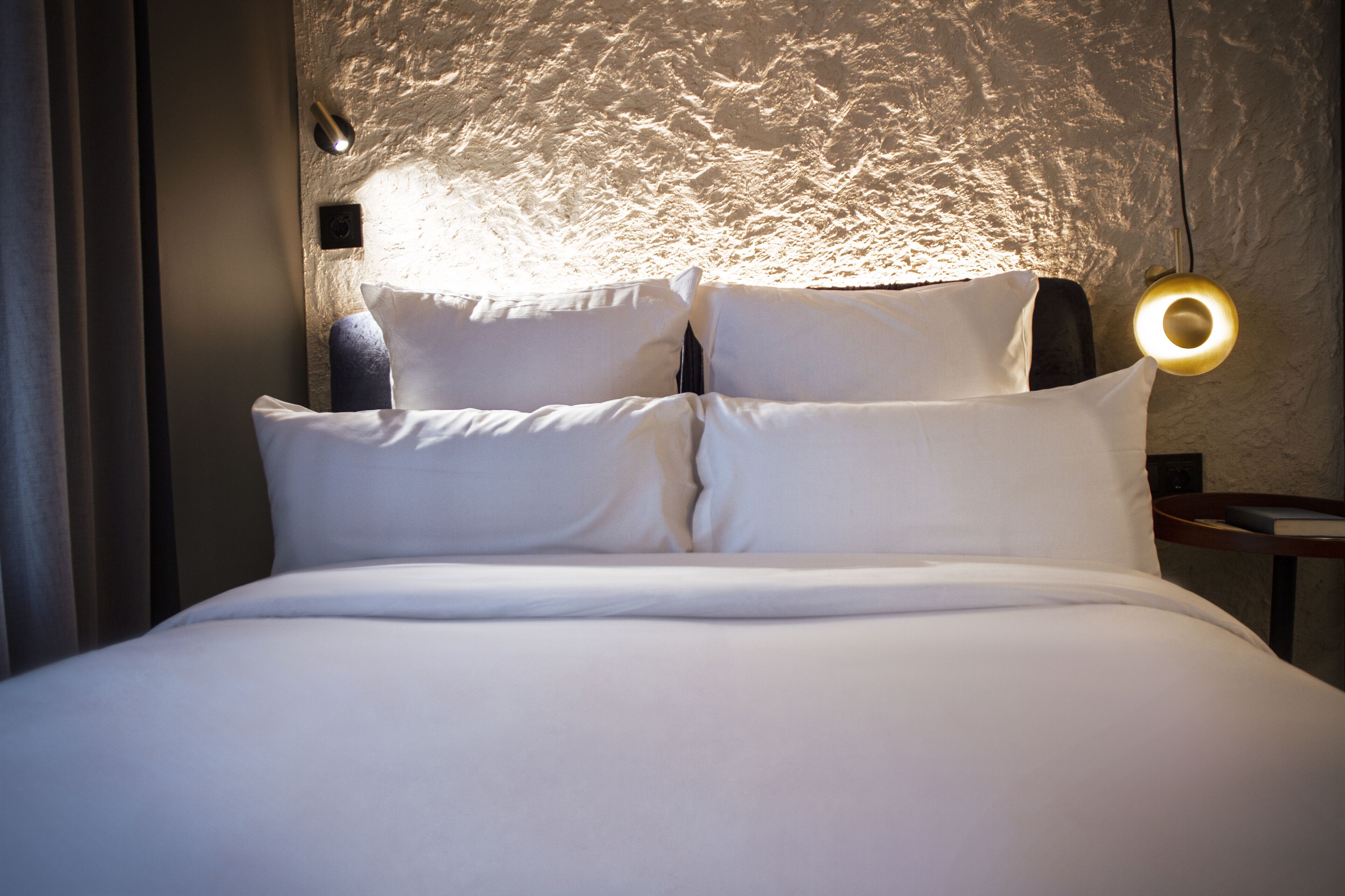 A neatly made bed with white linens and pillows, a textured wall behind, and a round wall-mounted light fixture.