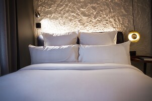 Un letto ben rifatto con lenzuola e cuscini bianchi, una parete strutturata sullo sfondo e un lampadario rotondo fissato al muro.