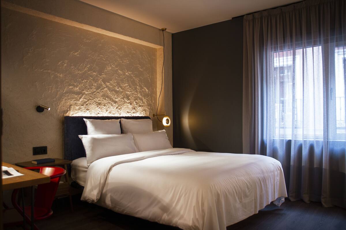 Un lit bien fait, avec du linge de lit blanc, dans une pièce dont un mur est texturé, un bureau avec une chaise rouge et une fenêtre ornée de rideaux légers.