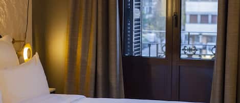 A hotel room with a bed, curtains, a window with blinds, and a view of buildings outside.