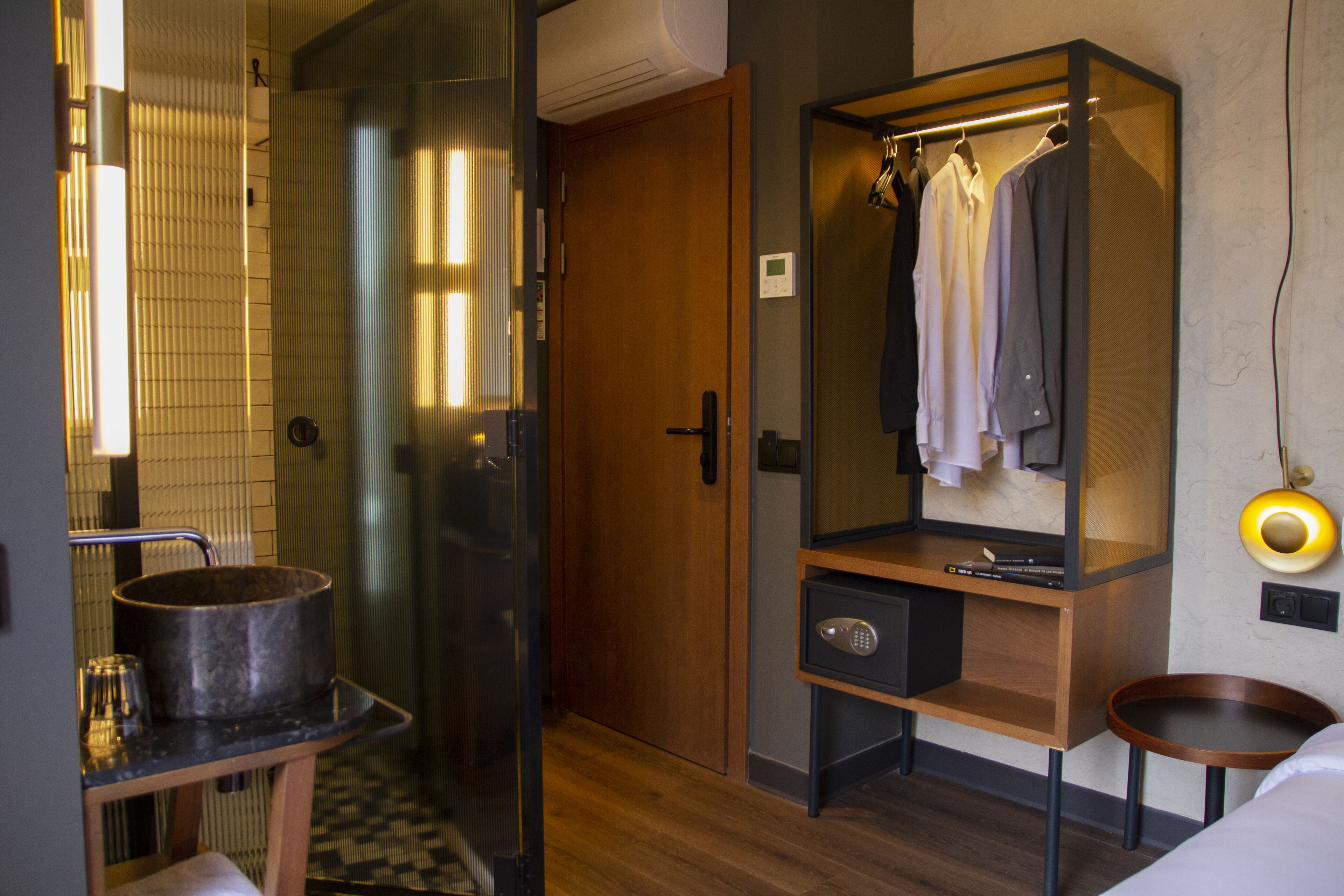A hotel room with a wooden wardrobe, a mirror, a small table with a pot, and a bedside table with a lamp.
