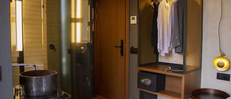 A hotel room with a wooden wardrobe, a mirror, a small table with a pot, and a bedside table with a lamp.