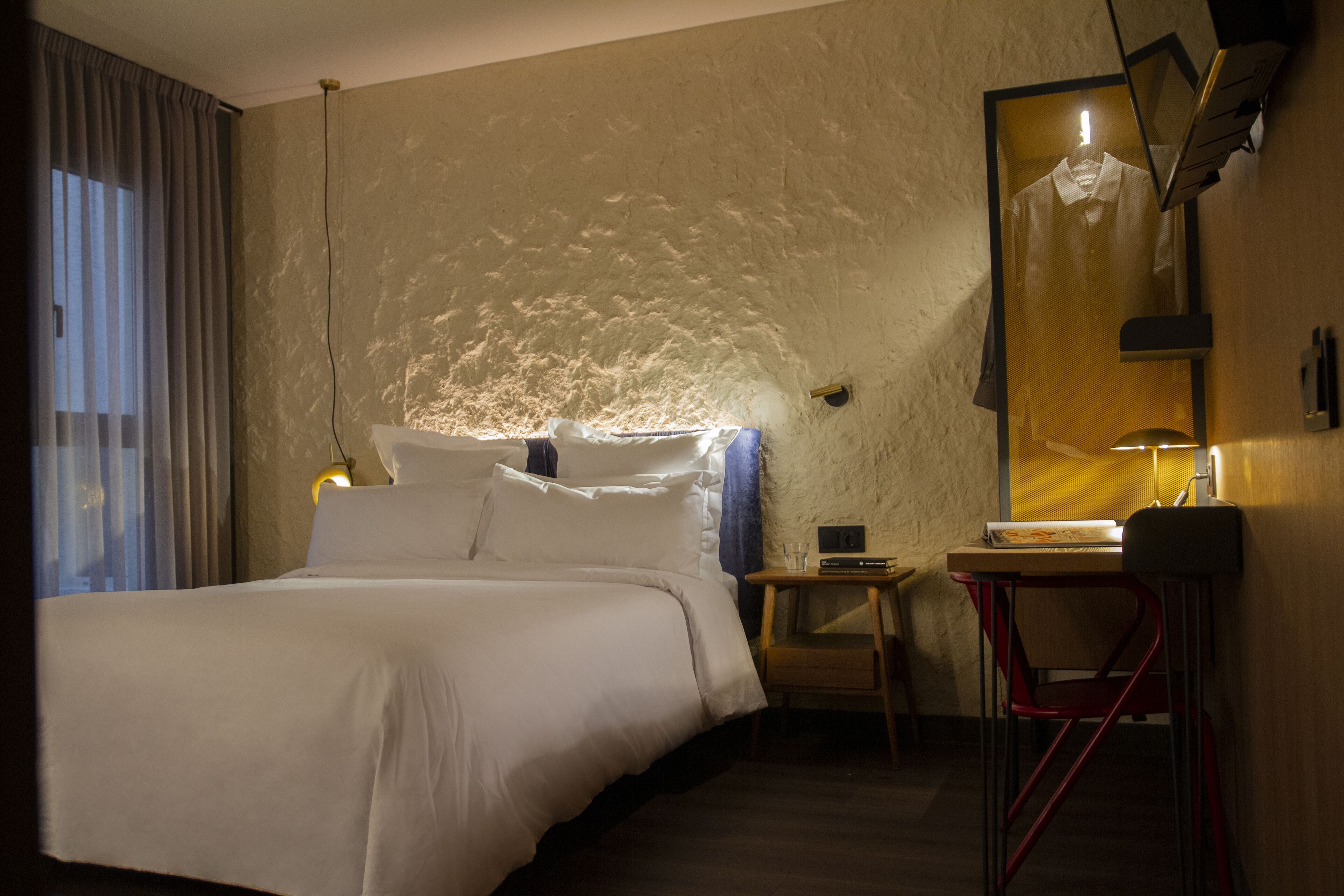 A hotel room with a bed, a desk, a chair, a mirror, and a window with curtains.