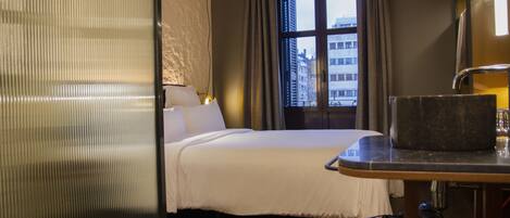 A hotel room with a bed, a desk with a lamp, a window with curtains, and a view of buildings outside.