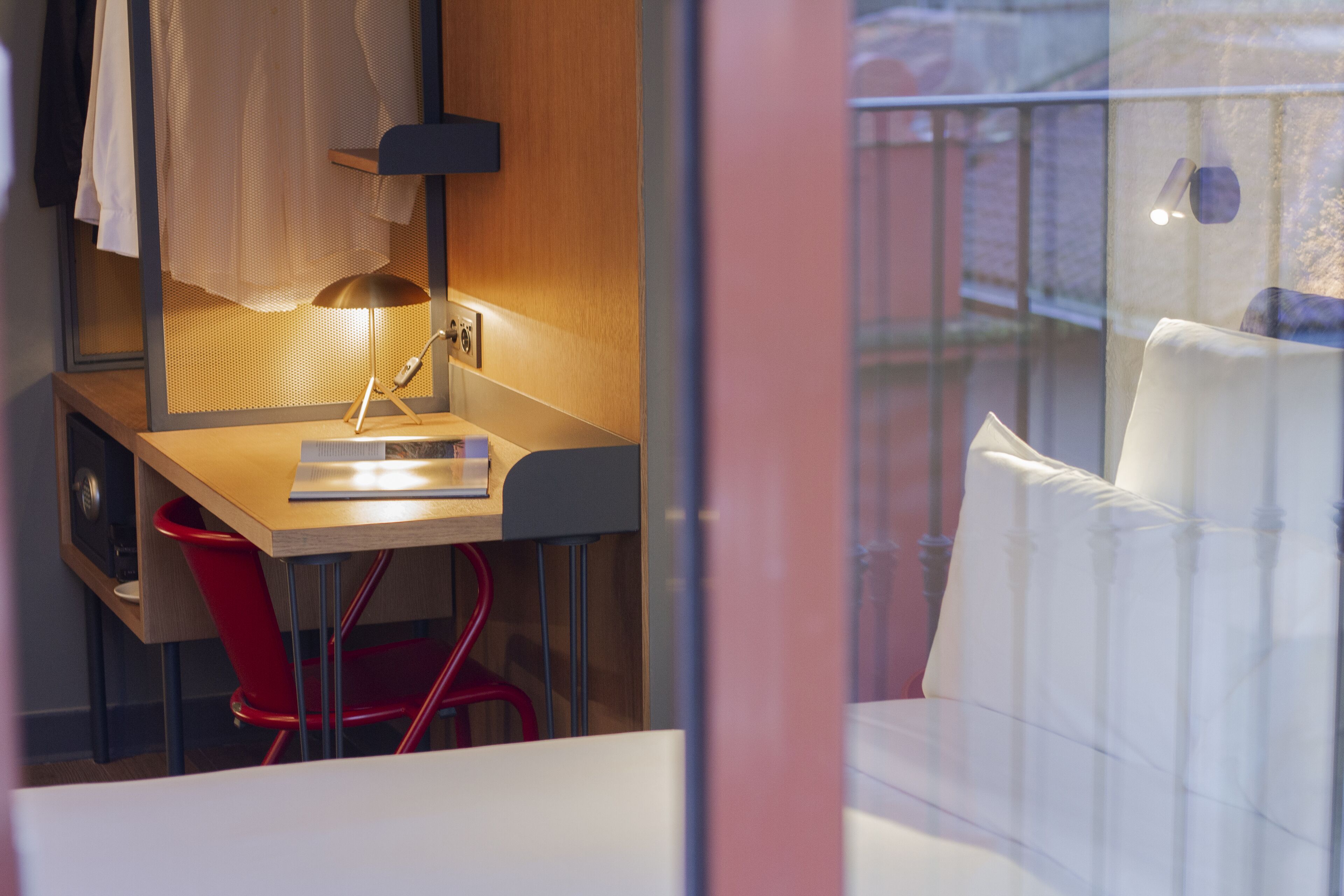 A hotel room with a desk, a red chair, a white sofa, and a window with a view of a balcony.