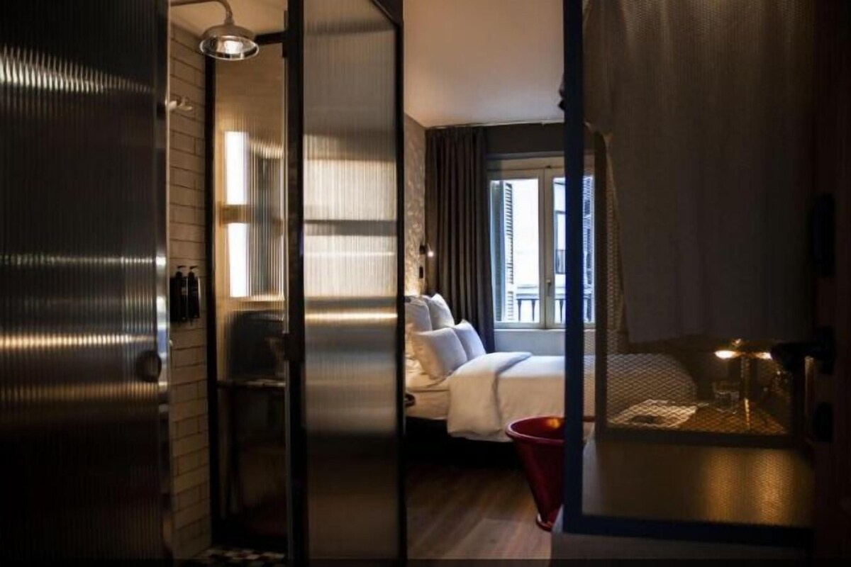 A hotel room with a bed, a small table, a red bucket, and a window with curtains.
