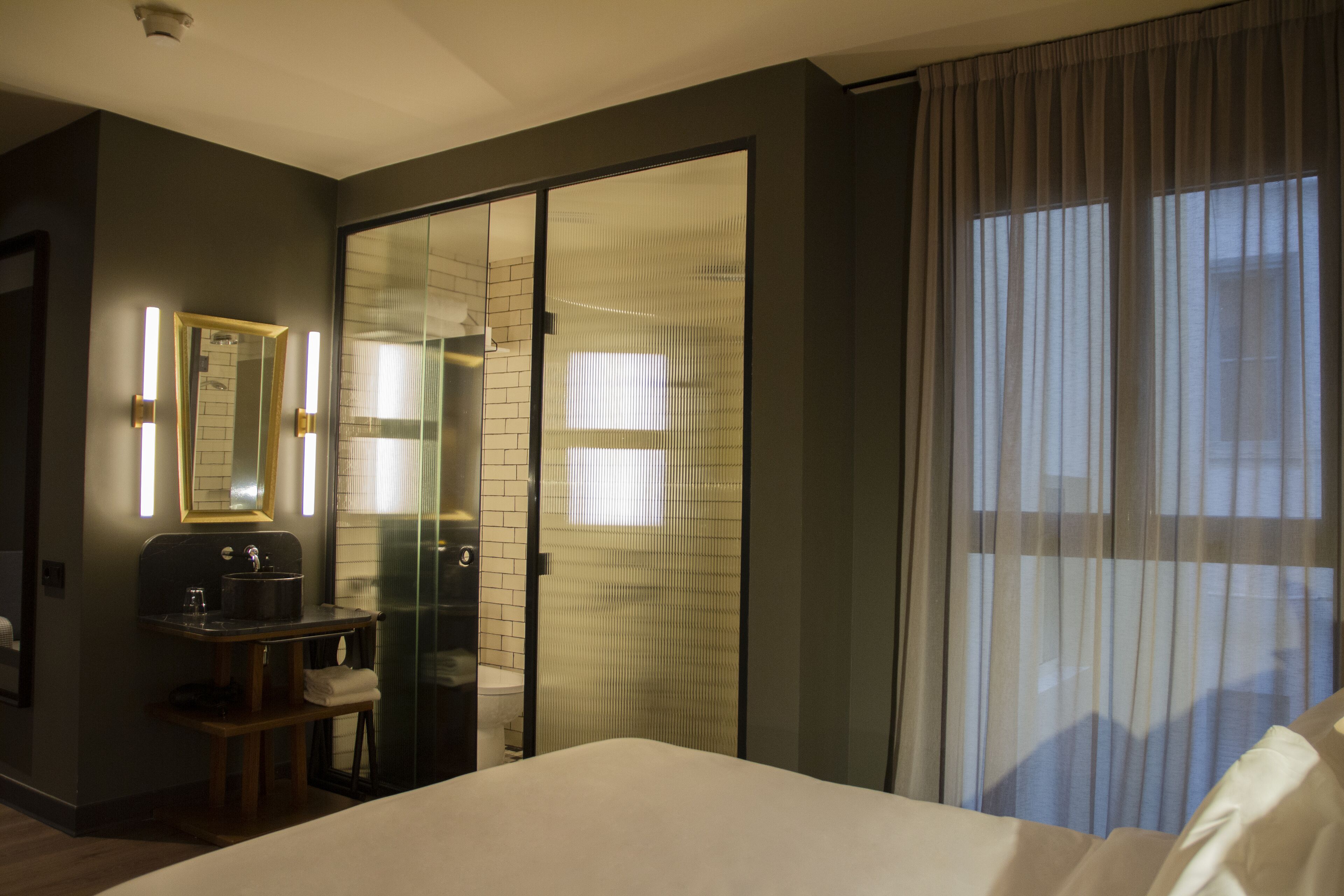 Quarto de hotel com uma cama, uma pequena escrivaninha com espelho, uma janela com cortinas e uma porta de vidro deslizante que dá para um banheiro.