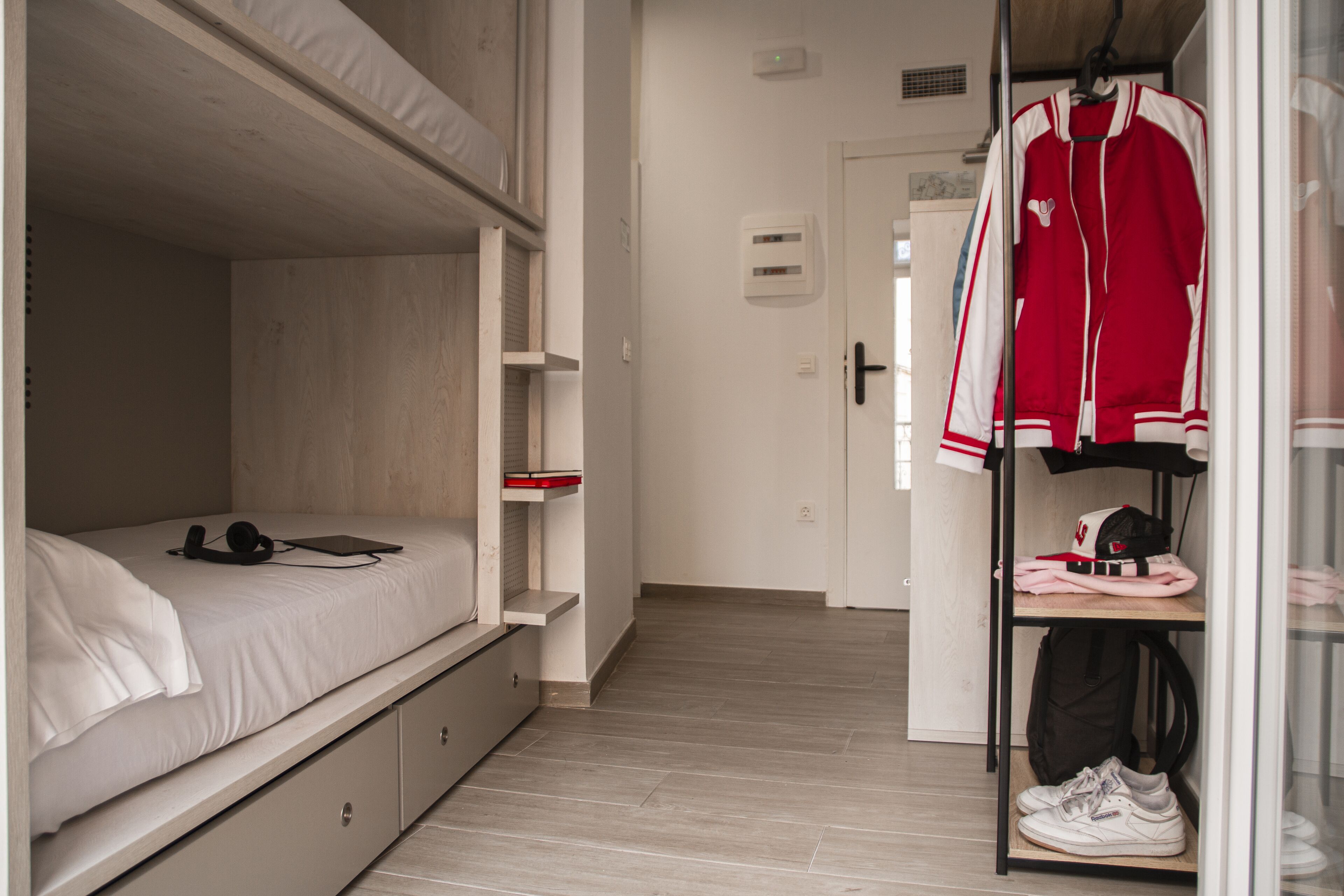 Quarto com cama beliche, mesa de cabeceira com fones de ouvido e um tablet, guarda-roupa com roupas e sapatos, e porta que dá para outro cômodo.