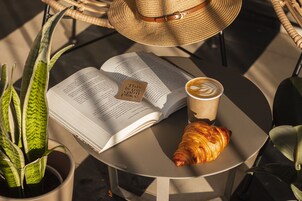Un libro, un croissant, una taza de café y un sombrero de paja sobre una mesa redonda.