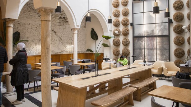 A spacious indoor area with wooden tables and benches, a brick wall, and a large window with a view. - Select Tetuán (Seville)