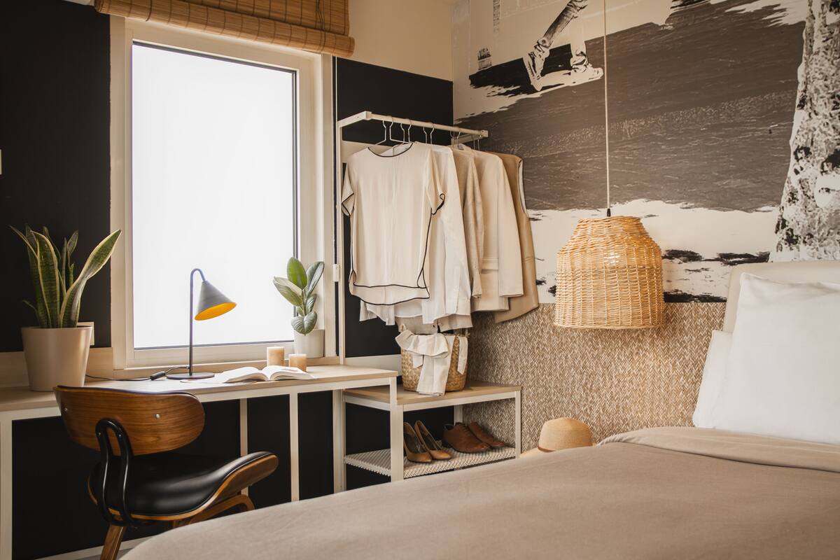 A bedroom with a bed, a desk with a chair, a wardrobe, a hanging lamp, and a potted plant.