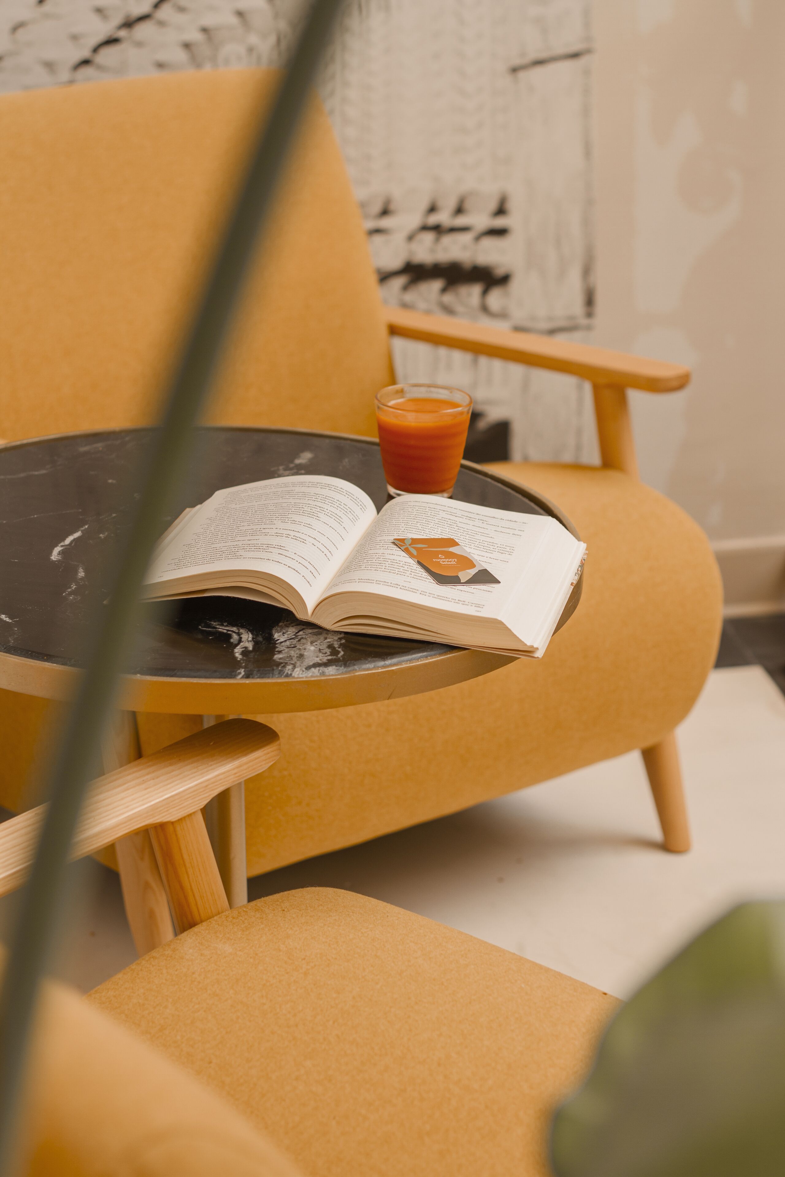 A yellow armchair with a book and a glass of orange juice on a small round table.