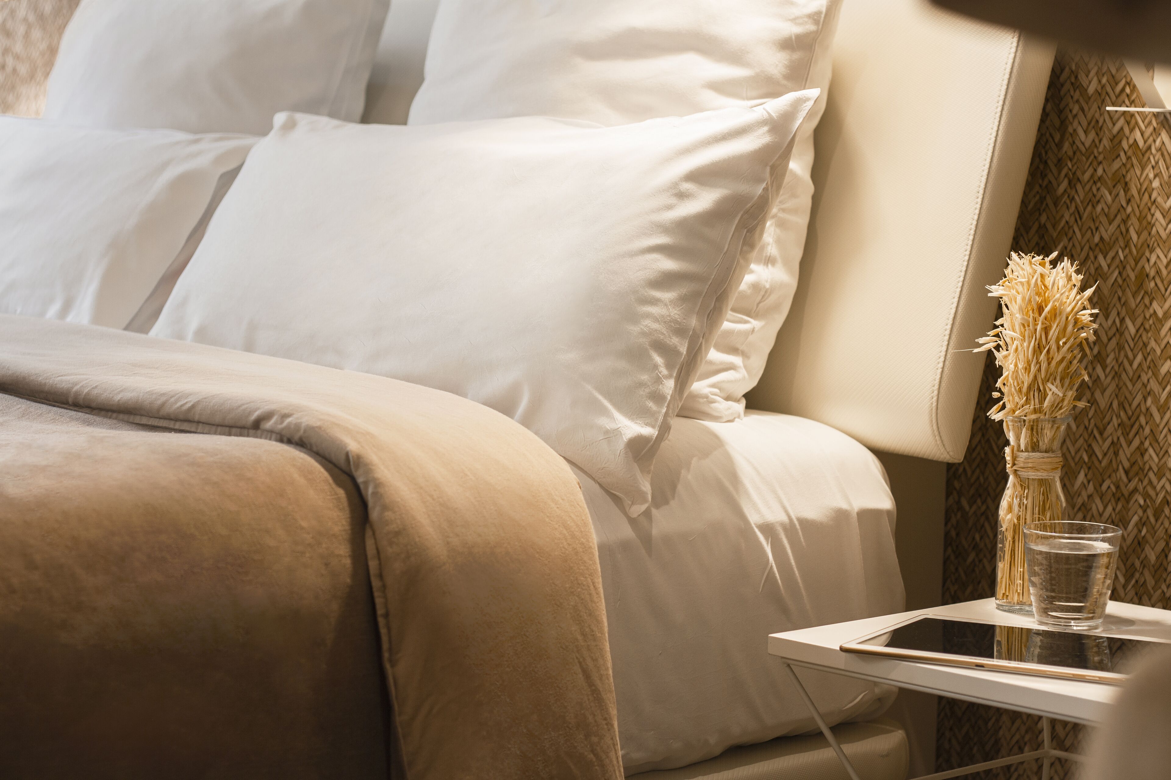 Una cama bien hecha con sábanas blancas, una mesita de noche con un vaso de agua y una tablet, y un florero con flores secas.