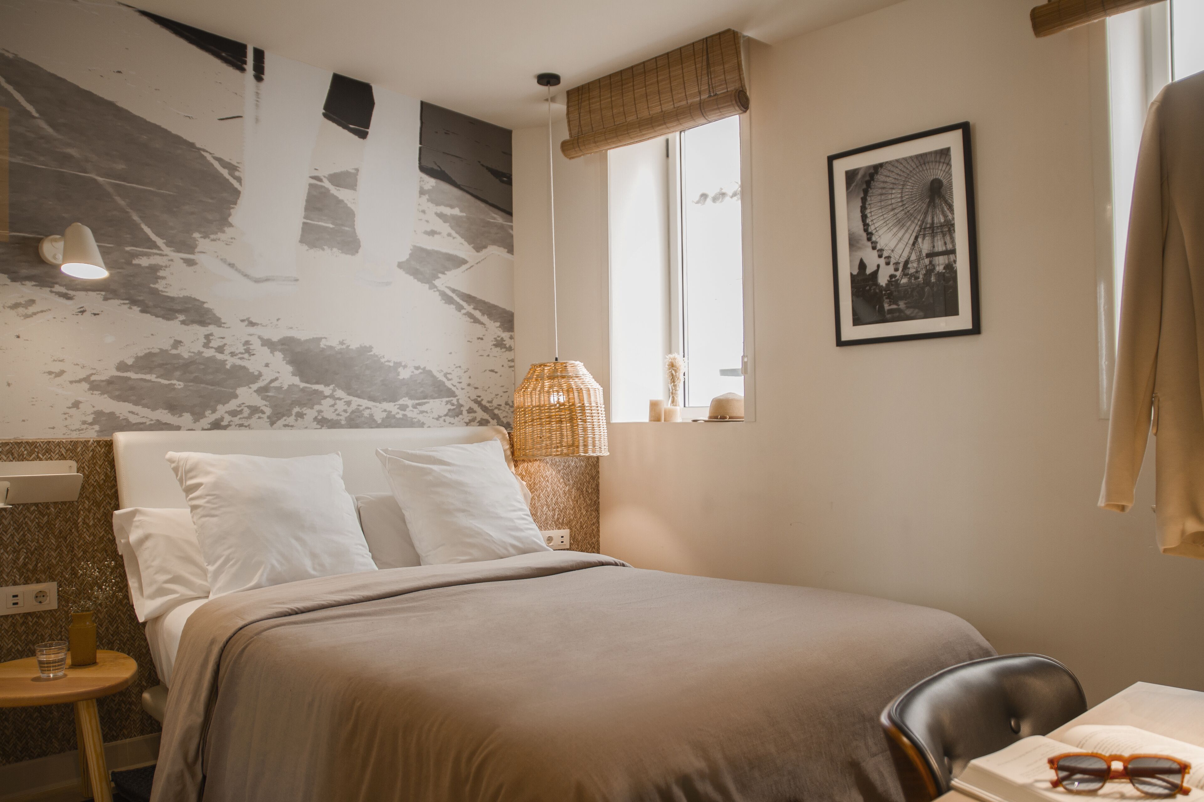 A hotel room with a bed, a small table with a lamp, a chair, a window with a view of a Ferris wheel, and a wall with a textured design.
