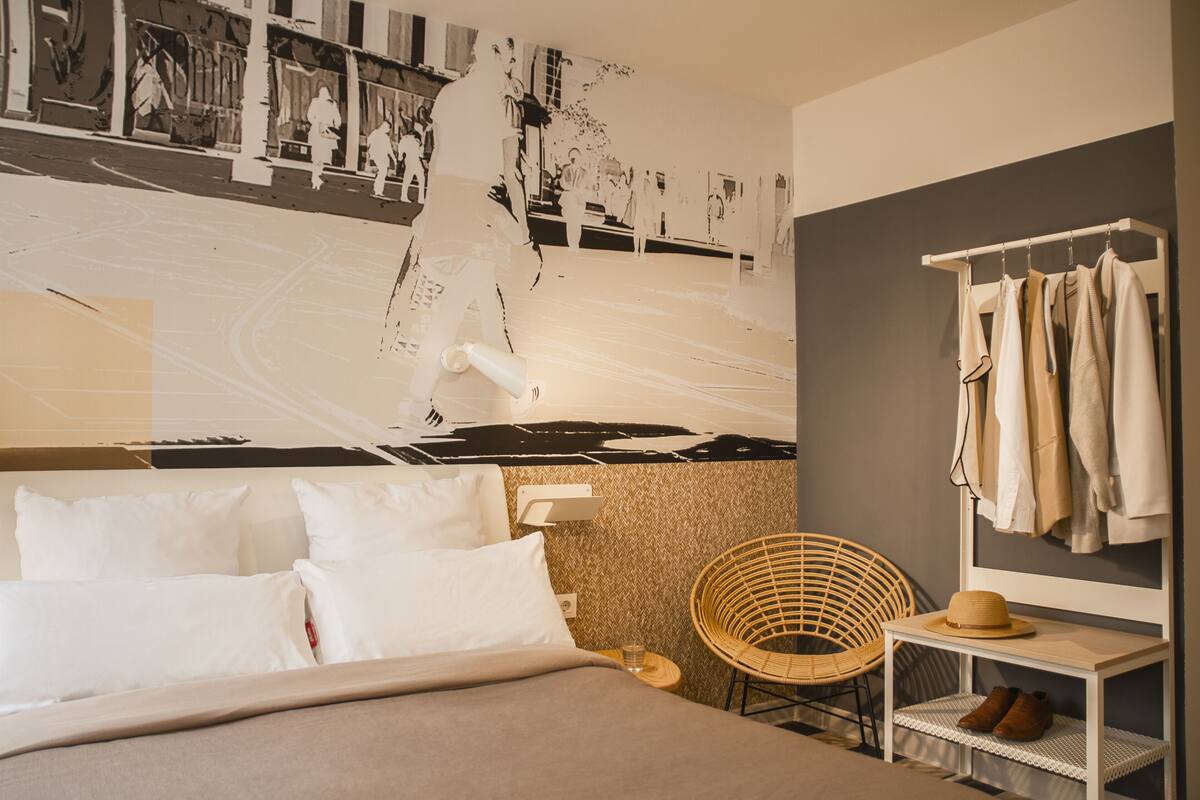A hotel room with a bed, a chair, a coat rack, and a wall with a large black and white photo.
