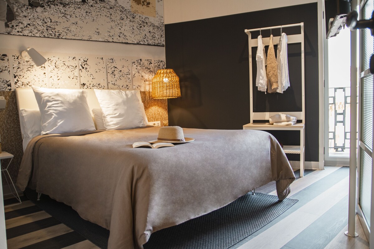 Una cama bien hecha con un libro y un sombrero, una lámpara de noche y un perchero para toallas.