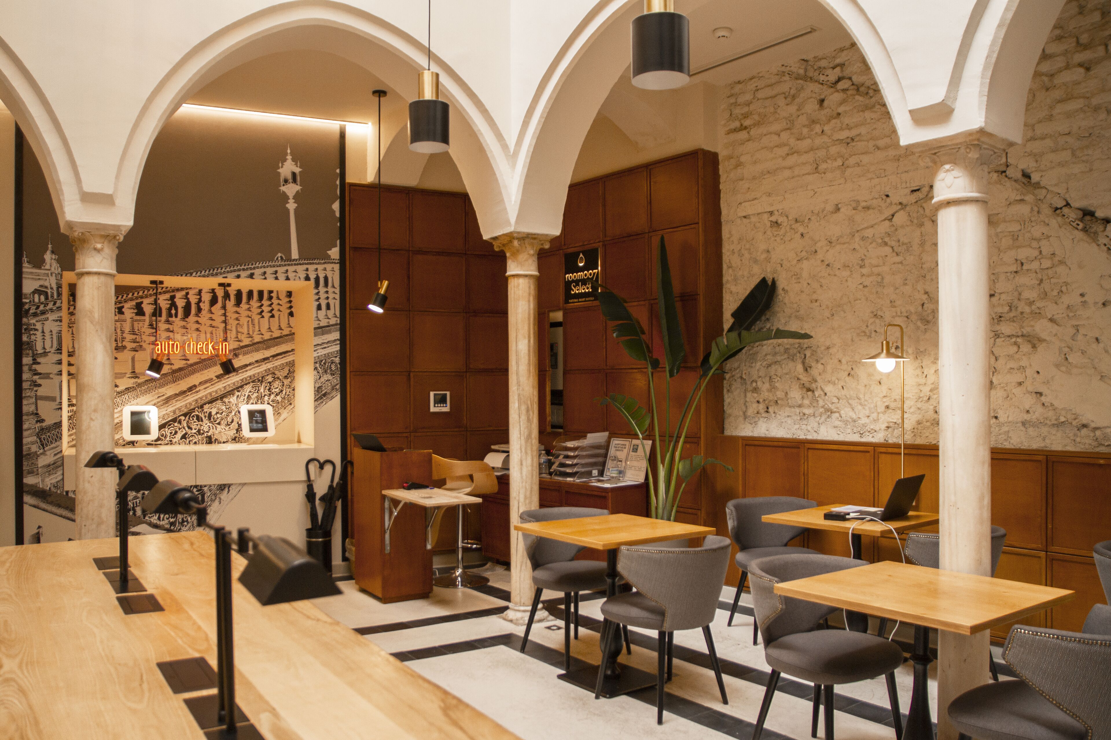 A modern hotel lobby with a wooden reception desk, multiple seating areas, and a staircase in the background.