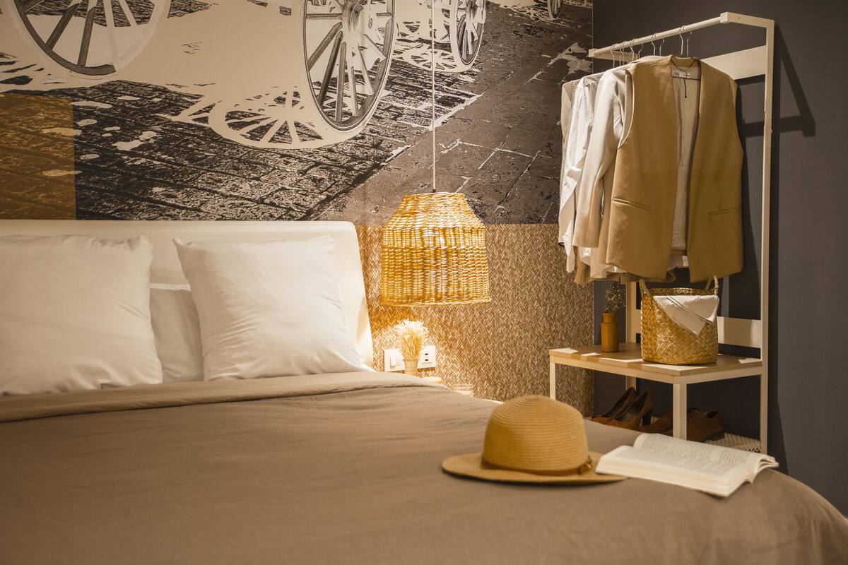 A neatly made bed with white linens, a wicker lamp, a hat, and a clothing rack with hanging clothes.