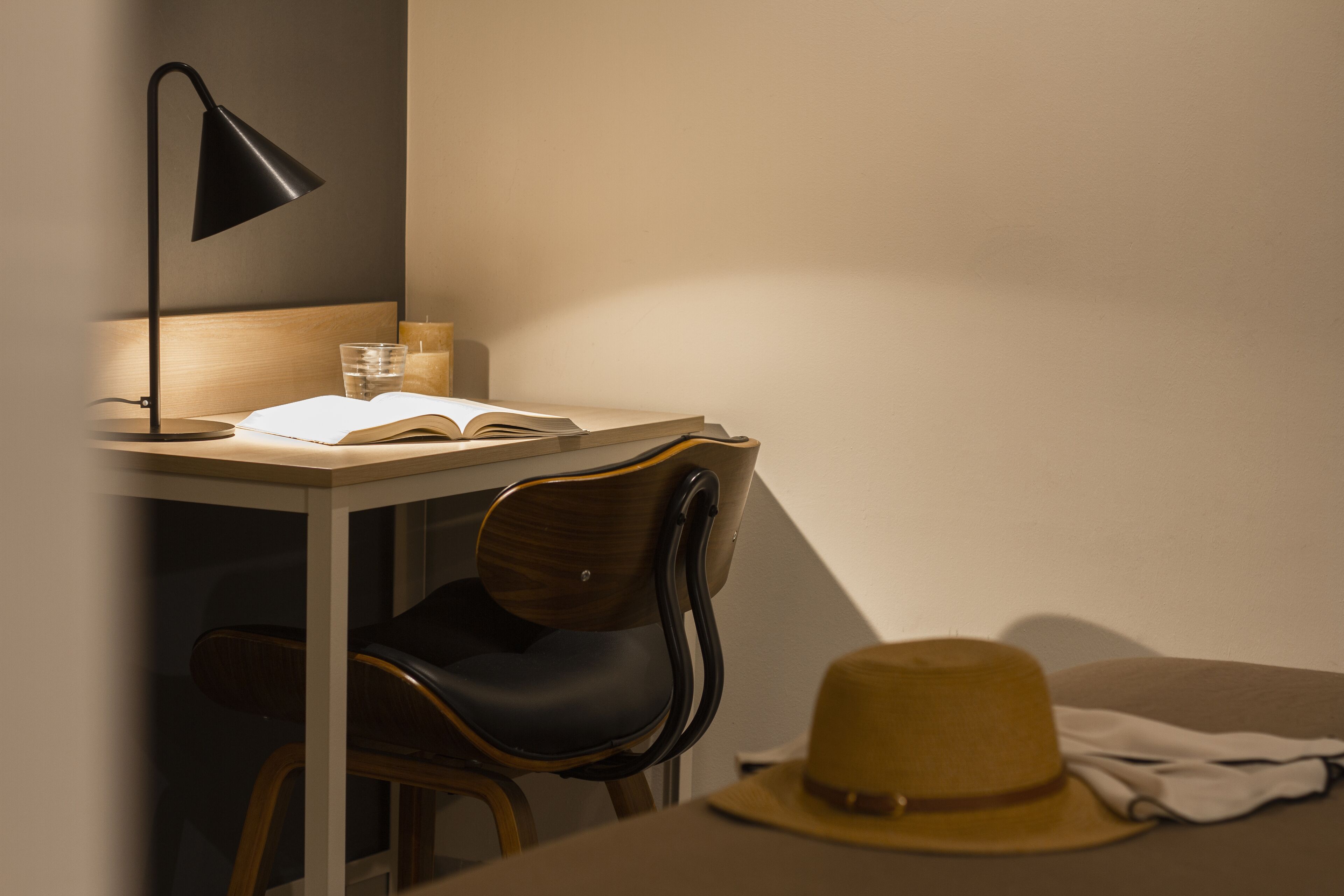 A study area with a desk, chair, lamp, open book, glass of water, and a hat.