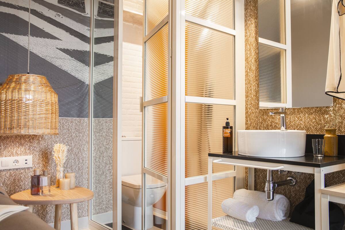 A modern bathroom with a glass partition, a round wooden table with toiletries, a white sink, and a mirror above it.
